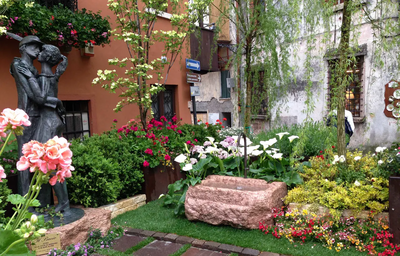 Aiuola d'autore attorno a una scultura in bronzo, con gerani, calle e bosso su fondo di prato e pietra